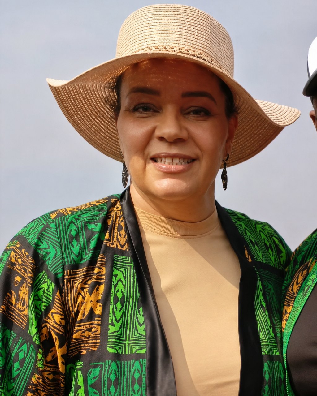 MIO smiling outdoors in a sun hat and green patterned jacket